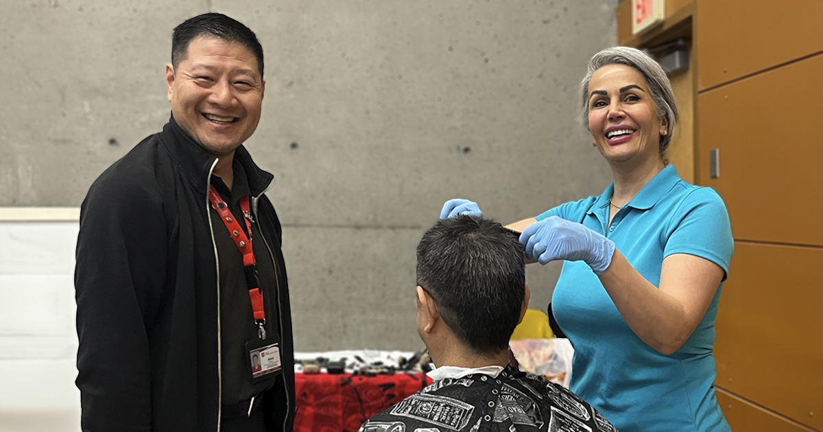 Man and woman stand together smiling, woman is cutting a third person's hair